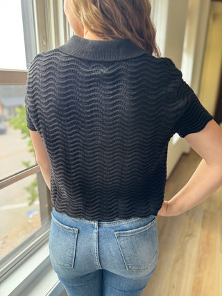 Person wearing a dark blue textured shirt and light blue jeans standing by a window.