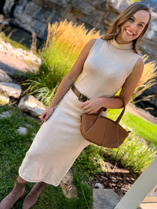A woman wearing a sleeveless cream knit mock-neck midi dress with a brown belt, holding a brown leather handbag, standing outdoors in a garden setting.