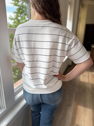 Person wearing a white and black striped shirt and blue jeans standing by a window.