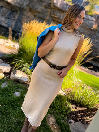 A woman wearing a sleeveless beige knit midi dress with a brown belt, standing outdoors in a garden setting with a blue jacket draped over her shoulder.