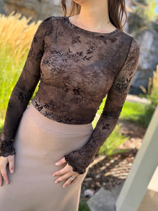 A woman wearing a sheer dark brown floral lace long-sleeve crop top paired with a beige knit skirt, photographed outdoors in a garden setting.