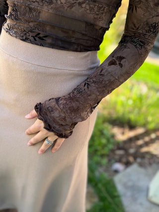A close-up of a person wearing sheer dark brown floral lace long gloves over a beige ribbed skirt, with a blurred outdoor garden background.