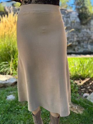 A beige knit midi skirt worn outdoors on a grassy lawn with rocks and greenery in the background.