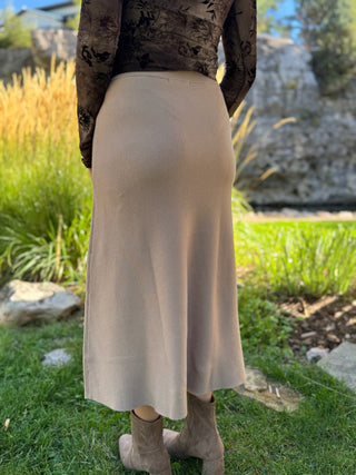 A person standing outdoors on grass wearing a light beige midi skirt, a dark patterned long-sleeve top, and taupe ankle boots, with a garden and rock wall in the background.