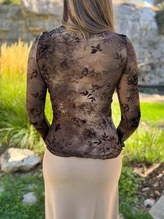 A woman viewed from behind wearing a sheer brown floral lace long-sleeve top over a light beige skirt, standing outdoors in a garden setting.