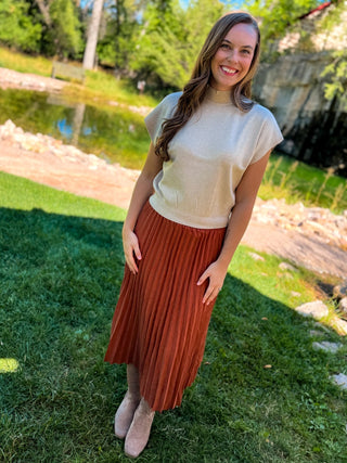 A woman standing outdoors on grass near a pond, wearing a cream short-sleeve knit top and a rust-colored pleated midi skirt with beige ankle boots.