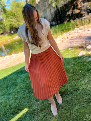 A woman standing on a grassy lawn outdoors, wearing a cream short-sleeve top and a rust-orange pleated midi skirt, paired with light pink pointed-toe heels.