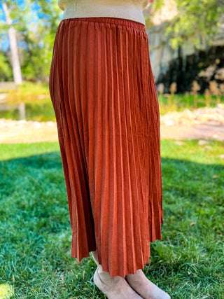 A rust-orange pleated midi skirt worn outdoors on a grassy lawn with trees in the background.