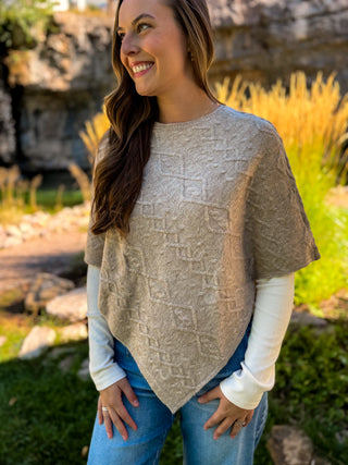 A woman wearing a beige textured knit poncho sweater over a white long-sleeve shirt and blue jeans, standing outdoors in a garden setting.