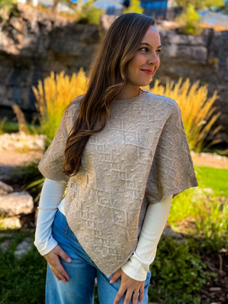 A woman wearing a beige textured knit poncho over a white long-sleeve shirt and blue jeans, standing outdoors in a garden setting with rocks and greenery.