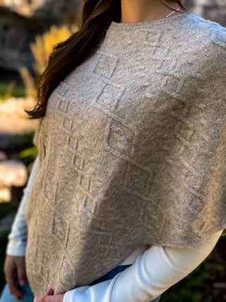 A close-up of a person wearing a chunky knit poncho sweater in a warm taupe-beige color with a raised geometric diamond pattern. The poncho is layered over a white long-sleeve shirt, and the background is a softly blurred outdoor garden setting.