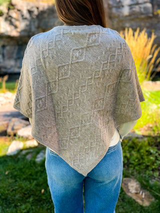A woman viewed from behind wearing a light taupe knit poncho with an embossed geometric diamond pattern, paired with blue jeans in an outdoor garden setting.
