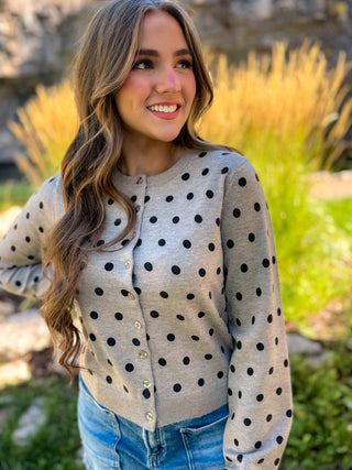 A woman wearing a light gray long-sleeve cardigan with black polka dots and gold buttons, paired with blue jeans, photographed outdoors in front of a blurred garden background.