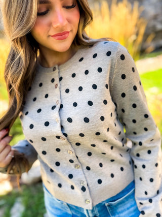 A woman wearing a light gray long-sleeve button-up cardigan with black polka dots, paired with blue jeans, photographed outdoors with a blurred natural background.