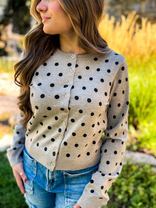 A woman wearing a beige cardigan with black polka dots and light-colored buttons, paired with blue jeans, photographed outdoors in a garden setting.
