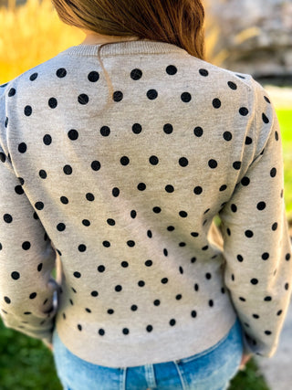 A person viewed from behind wearing a light heather gray long-sleeve sweater with black polka dots, paired with blue jeans, outdoors with blurred green grass and warm sunlight in the background.