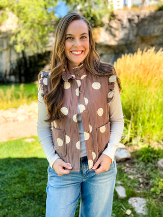 A smiling woman wearing a brown sleeveless vest with large cream polka dots over a beige long-sleeve shirt and blue jeans, standing outdoors in a garden setting.