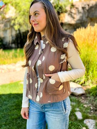 A woman wearing a brown sleeveless vest with large cream polka dots over a beige long-sleeve shirt and blue jeans, standing outdoors in a grassy garden setting.