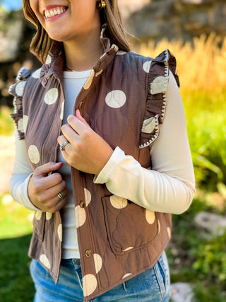 A woman wearing a brown sleeveless quilted vest with beige polka dots and ruffled trim, layered over a cream long-sleeve top and blue jeans, photographed outdoors with a blurred natural background.