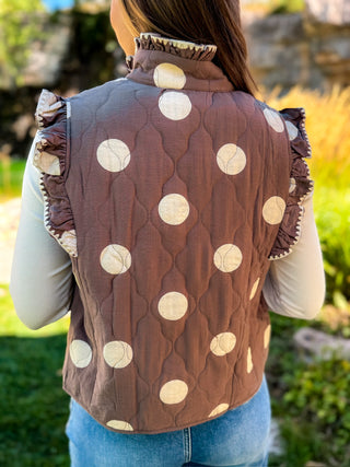 A person viewed from the back wearing a brown quilted sleeveless vest with large cream polka dots and ruffled trim, over a light long-sleeve top and blue jeans, outdoors on a grassy area.