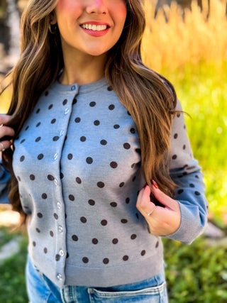 A woman wearing a light blue-gray cardigan with dark brown polka dots, paired with blue jeans, photographed outdoors with a blurred natural background.