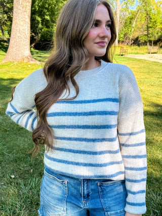 A woman wearing a cream knit sweater with blue horizontal stripes and blue jeans, standing outdoors in a grassy park area with trees in the background.