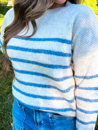 A person wearing a fuzzy knit sweater with horizontal blue and off-white stripes, paired with blue jeans, photographed outdoors on grass.