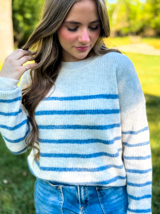 A woman wearing a white and blue horizontally striped knit sweater with blue jeans, standing outdoors in a grassy area.