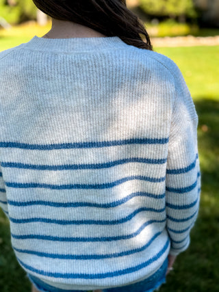 A person viewed from behind wearing a light gray knit sweater with horizontal blue stripes, standing outdoors on a grassy lawn.