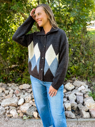 A woman wearing a dark charcoal knit cardigan with a blue and cream argyle pattern, paired with light blue jeans, standing outdoors in front of a background of green foliage and rocks.