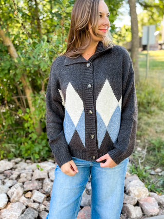 A woman wearing a dark charcoal gray knit cardigan with a large argyle pattern in cream, light blue, and dark gray, paired with blue jeans, standing outdoors on a bed of rocks with green foliage in the background.