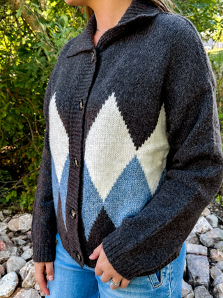 A person wearing a dark charcoal knit cardigan with an argyle pattern in cream, light blue, and dark brown, paired with blue jeans, outdoors on a rocky path with green foliage in the background.