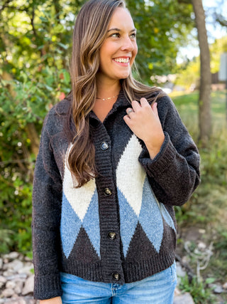 A woman wearing a dark charcoal gray knit cardigan with a large argyle pattern in cream, light blue, and dark gray, paired with blue jeans, standing outdoors in a garden setting.