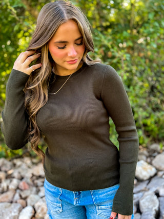 A woman wearing a ribbed long-sleeve crewneck sweater in dark olive brown, paired with blue jeans, standing outdoors in front of a blurred green foliage background.
