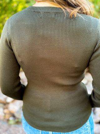 A close-up back view of a person wearing a ribbed long-sleeve top in olive green, paired with blue jeans, outdoors with a blurred natural background.