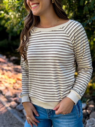 A woman wearing a cream and brown horizontally striped long-sleeve top with a boat neckline, paired with blue jeans, photographed outdoors in front of a blurred garden background.