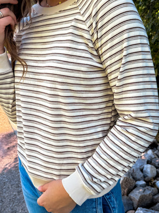 A person wearing a cream-colored long-sleeve crewneck sweater with thin brown horizontal stripes, paired with blue jeans, shown in an outdoor setting.