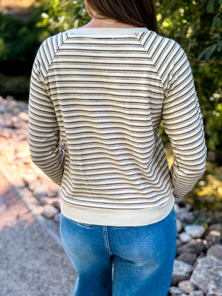 A woman viewed from behind wearing a cream and brown horizontally striped long-sleeve top with a wide boat neckline, paired with blue jeans, outdoors on a paved path with greenery in the background.