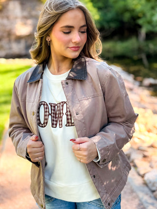 A woman wearing a taupe-brown utility jacket with a dark brown plaid collar, layered over a cream graphic sweatshirt and blue jeans, standing outdoors on a stone path.