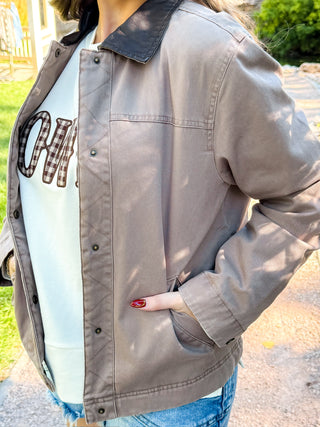 A person wearing a taupe-gray utility jacket with a black collar, layered over a white graphic t-shirt and blue jeans, photographed outdoors in natural light.