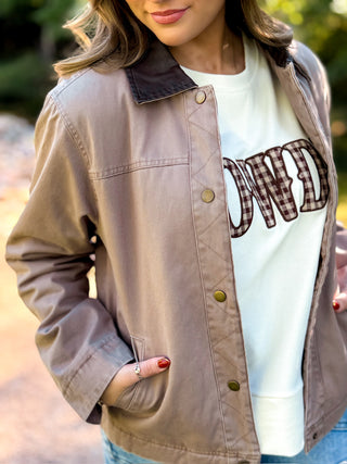 A woman wearing a taupe-brown utility jacket over a white graphic t-shirt with blue jeans, photographed outdoors with a blurred natural background.
