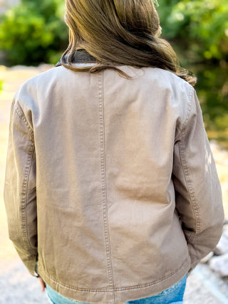 A person viewed from behind wearing a light tan canvas utility jacket, with green foliage and a stone path blurred in the background.