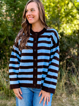 A woman wearing a blue and dark brown striped knit cardigan sweater with blue jeans, standing outdoors in front of a blurred green foliage background.
