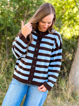 A woman wearing a brown and light blue striped button-up cardigan sweater and blue jeans, standing outdoors in front of a blurred green background.