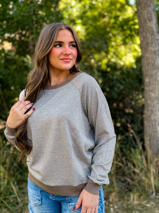 A woman wearing a light gray and brown horizontally striped long-sleeve sweatshirt with solid brown ribbed trim, standing outdoors in front of a blurred green forest background.