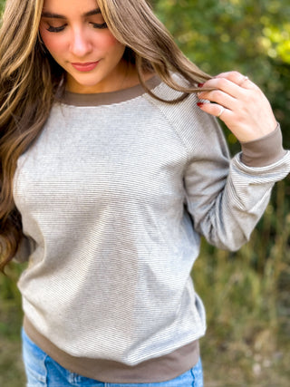A woman wearing a light gray and brown ribbed long-sleeve top, shown from the chest down in an outdoor setting with blurred green foliage in the background.
