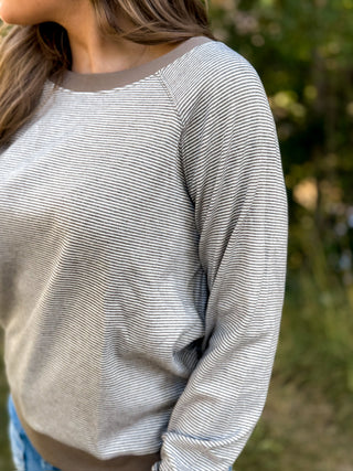 A close-up of a woman wearing a long-sleeve crewneck sweater with fine horizontal gray and white stripes, featuring solid taupe-brown ribbed trim at the collar and hem, set against a blurred outdoor background.