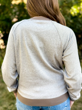A person viewed from behind wearing a light beige and gray horizontally striped long-sleeve top with a solid brown ribbed collar, hem, and cuffs, paired with blue jeans, set against a blurred green outdoor background.
