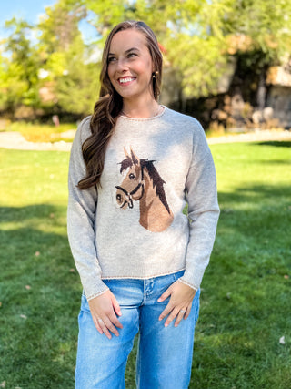 A woman standing outdoors on a grassy lawn wearing a light gray knit sweater with a large horse face graphic on the front, paired with blue jeans.