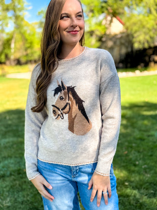 A woman wearing a light heather gray knit sweater with a detailed horse head graphic on the front, paired with blue jeans, standing outdoors on a grassy lawn.